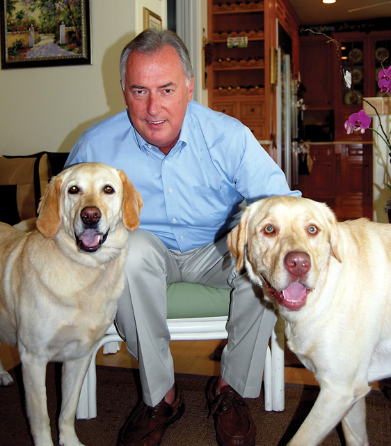 Gerry Weber, Fast-Fix CEO, pictured embracing his 2 rescued golden labs. He is wearing a blue shirt and cream color pants in a living room environment.