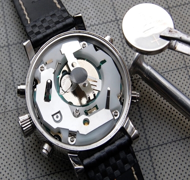 Back of a watch opened, showing the gears, and a battery being approached to it with tweezers.