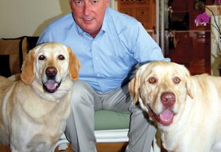 Gerry Weber, Fast-Fix CEO, pictured embracing his 2 rescued golden labs. He is wearing a blue shirt and cream color pants in a living room environment.